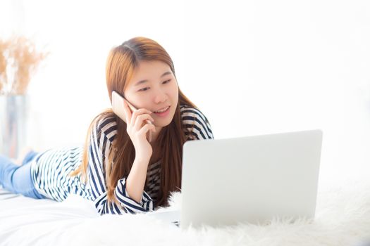 Beautiful asian young woman lying on bed using laptop and talking mobile smart phone at bedroom for leisure and relax, freelance with girl working notebook, communication concept.
