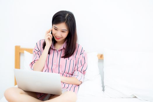 Portrait of beautiful asian young woman setting on bed using laptop computer and talk smart phone at bedroom for leisure and relax, freelance with girl working notebook and calling telephone, communication and lifestyle concept.