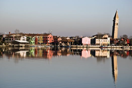 The colorful village of Burano, with its characteristic leaning bell tower, is reflected in the waters of the Venetian lagoon.