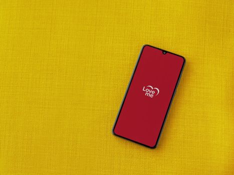 Lod, Israel - July 8, 2020: Loveme app launch screen with logo on the display of a black mobile smartphone on a yellow fabric background. Top view flat lay with copy space.