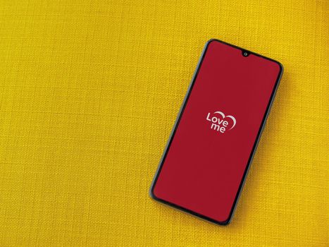 Lod, Israel - July 8, 2020: Loveme app launch screen with logo on the display of a black mobile smartphone on a yellow fabric background. Top view flat lay with copy space.