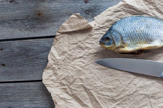 Salted Dry fish vobla with a sharp knife on crumpled craft paper on wooden background, delicious beer snack, close-up.