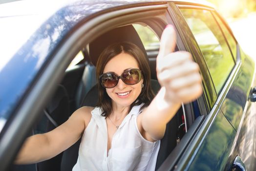 A smart looking middle-aged adult woman wearing sunglasses put her hand out the car window and raised her thumbs up.