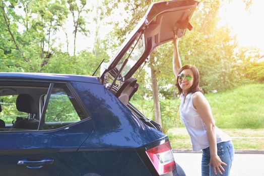 Smiling Caucasian woman in a white blouse and denim shorts putting her stuff bags into the car trunk