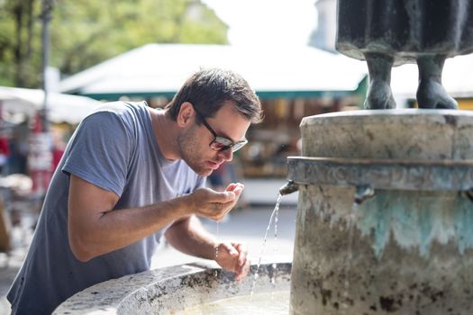 Thirsty young casual cucasian man drinking water from public city fountain on a hot summer day.