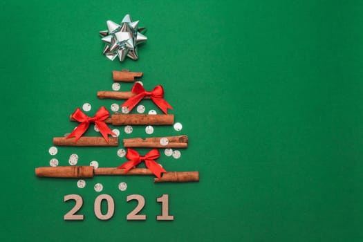 New year card 2021. Christmas tree made of cinnamon sticks with silver gift bow and small red bows on green background with silver confetti, copy space. Flat lay. template for design.