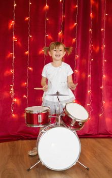 Little smiling girl plays on drums on red background with new year garlands.