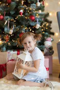 Pretty little child is sitting in front of christmas tree among garlands and presents..