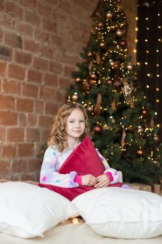 Cute little girl sits on the bed and huggs pillows with background of christmas tree behaind.