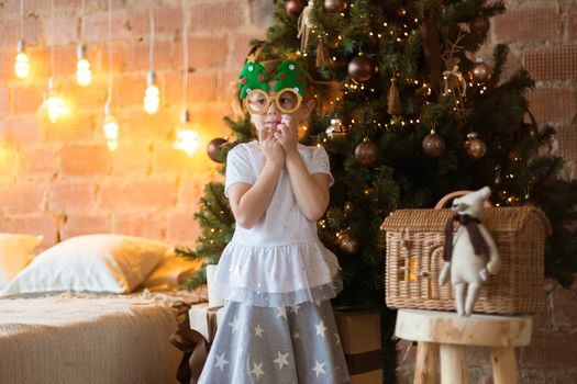 .Funny little girl in mask-glasses with a fir-tree on a christmas background in a loft interior