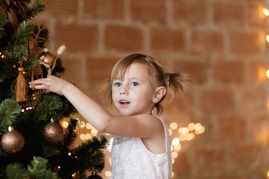 .Cute little girl in a chic dress hangs Christmas toys on the tree.