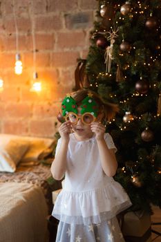 .Funny little girl in mask-glasses with a fir-tree on a christmas background in a loft interior