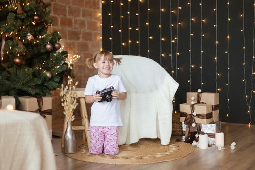 Adorable little girl in pajamas sits on the floor with camera in her hands near christmas tree.