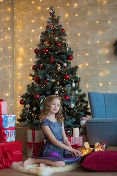 Pretty little child is sitting in front of christmas tree among garlands and presents..