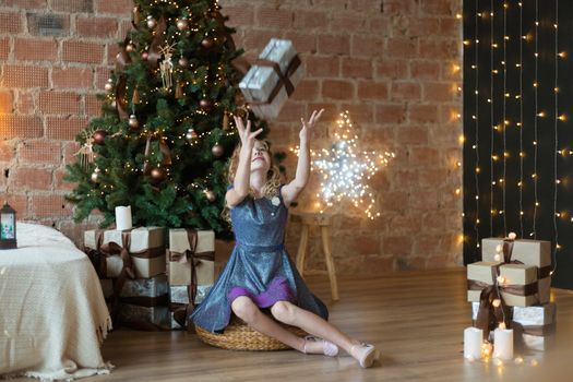 .Adorable little girl in a luxurious dress throws up a gift box near the Christmas tree.