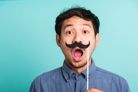 Portrait Asian happy handsome man posing he holding a funny mustache card or vintage fake moustaches on his mouth, studio shot isolated on blue background, Fathers day, November day concept