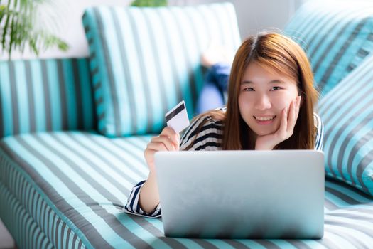 Beautiful of portrait young asian woman lying users credit card with laptop, Content girl shopping online and payment with notebook computer on sofa, lifestyle concept.