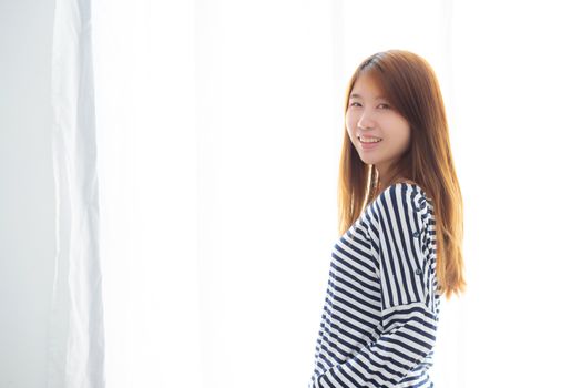 Portrait of beautiful young asian woman standing the window and smile while wake up with sunrise at morning, lifestyle and relax concept.