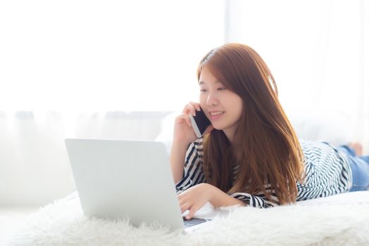 Beautiful asian young woman lying on bed using laptop and talking mobile smart phone at bedroom for leisure and relax, freelance with girl working notebook, communication concept.