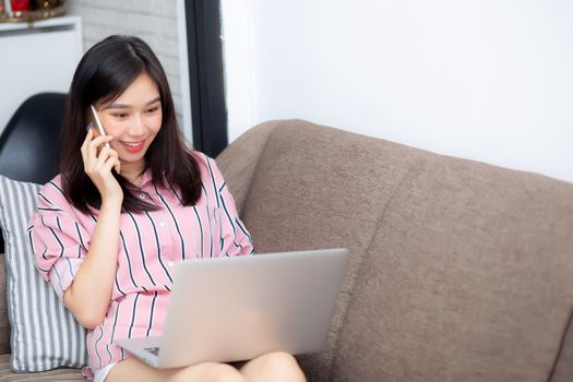 Beautiful asian young woman lying on bed using laptop and talking mobile smart phone on sofa at living room with satisfied for leisure and relax, freelance with girl working notebook, communication concept.