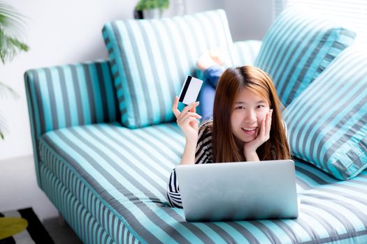 Beautiful of portrait young asian woman lying users credit card with laptop, Content girl shopping online and payment with notebook computer on sofa, lifestyle concept.