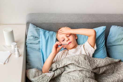 Sick little boy with high fever and headache laying in bed and holding thermometer in his mouth. Stay at home during corona virus epidemic if you feel sick