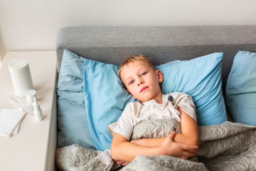 Sick little boy with high fever and headache laying in bed and holding thermometer. Stay at home during corona virus epidemic if you feel sick