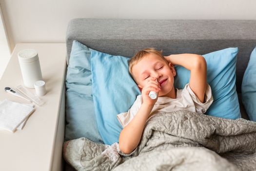 Sick little boy with high fever and headache laying in bed spraying medicine into his nose. Stay at home during corona virus epidemic if you feel sick