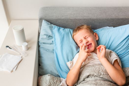 Sick little boy with high fever and headache laying in bed spraying medicine into his nose. Stay at home during corona virus epidemic if you feel sick
