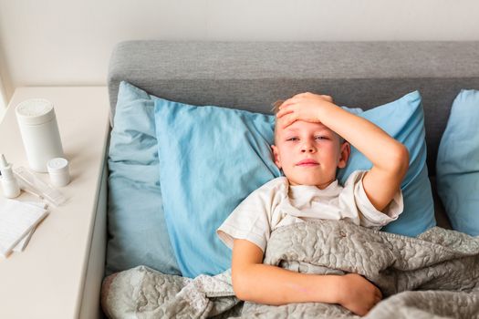 Sick little boy with high fever and headache laying in bed checking his forehead. Stay at home during corona virus epidemic if you feel sick