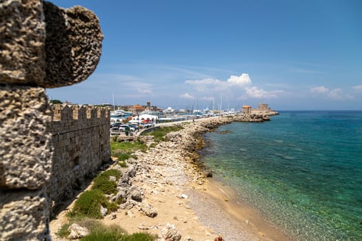 Coastline along the Mandraki  harbour in Rhodes city with gravel beach, crystal clear turquoise water, old windmills, Kastell Agios Nikolaos and lighthouse on Rhodes island, Greece 