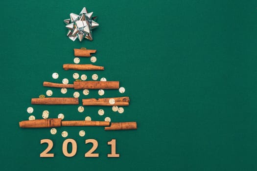 New Year card 2021. Christmas tree made of cinnamon sticks with silver gift bow on green background with silver confetti, copy space. Flat lay. template for design.