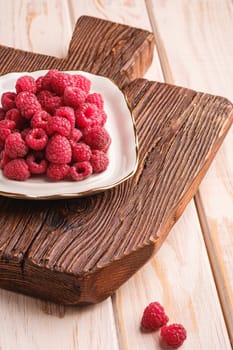 Raspberry fruits in plate on old teak cutting board, healthy pile of summer berries on wooden background, angle view