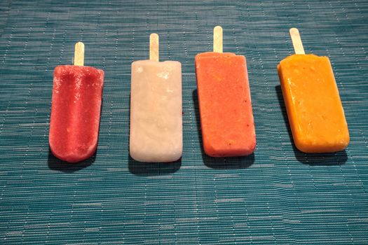 Fruit popsicles on an aqua blue background in summer including watermelon popsicle, tropical fruit popsicle, mango popsicle, and lime popsicle with fresh raspberries.
