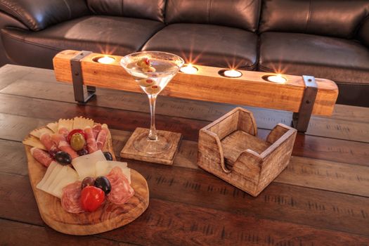 Martini with olives and a Charcuterie board on rustic wood with candles behind a spread of prosciutto panino, mozzarella cheese, Genoa salami, Fontina cheese and artisanal crackers.