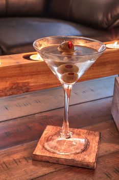 Dirty martini with green Pimento olive on a rustic wood coaster with rustic candles behind.