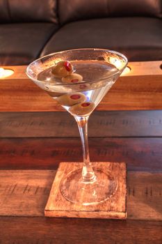 Dirty martini with green Pimento olive on a rustic wood coaster with rustic candles behind.