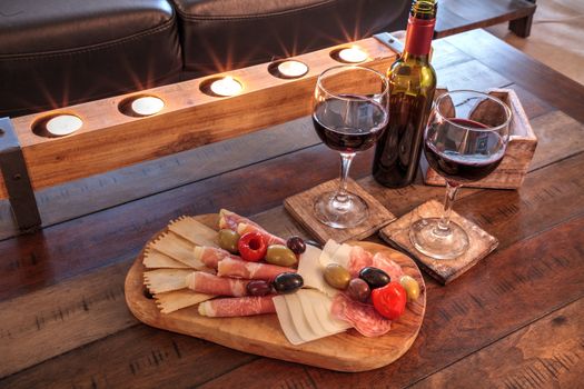 Red wine with Charcuterie board on rustic wood with candles behind a spread of prosciutto panino, mozzarella cheese, Genoa salami, Fontina cheese and artisanal crackers.