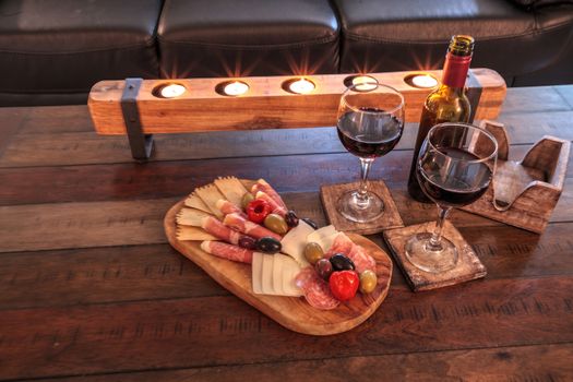 Red wine with Charcuterie board on rustic wood with candles behind a spread of prosciutto panino, mozzarella cheese, Genoa salami, Fontina cheese and artisanal crackers.