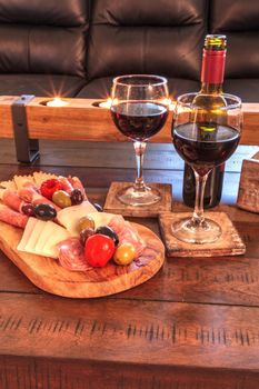 Red wine with Charcuterie board on rustic wood with candles behind a spread of prosciutto panino, mozzarella cheese, Genoa salami, Fontina cheese and artisanal crackers.