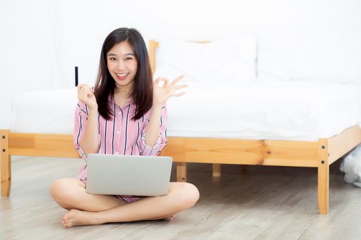 Beautiful portrait young asian woman sitting using credit card with laptop computer, content girl gesture ok and excited shopping online purchase with notebook in the bedroom, lifestyle concept.