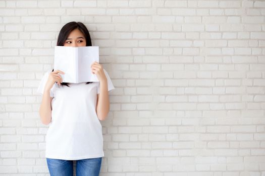 beautiful portrait young asian woman happy hiding behind open the book with cement or concrete background, girl standing reading for learning, education and knowledge concept.