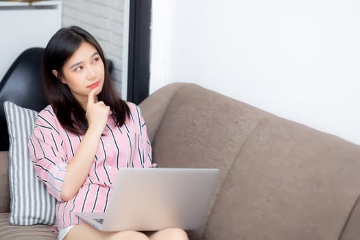 Beautiful of portrait asian young woman working online laptop and thinking sitting on couch, freelance girl using notebook computer on sofa, business and lifestyle concept.