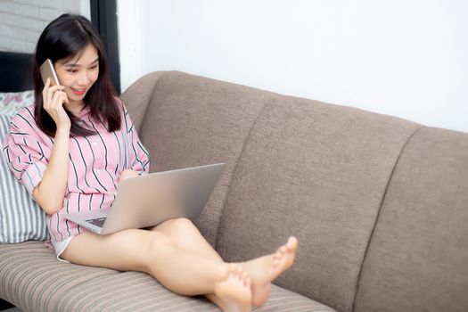 Beautiful asian young woman lying on bed using laptop and talking mobile smart phone on sofa at living room with satisfied for leisure and relax, freelance with girl working notebook, communication concept.