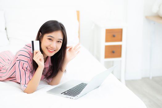 Beautiful of portrait young asian woman lying users credit card with laptop, content girl shopping online and payment with notebook computer on bed at bedroom, lifestyle concept.