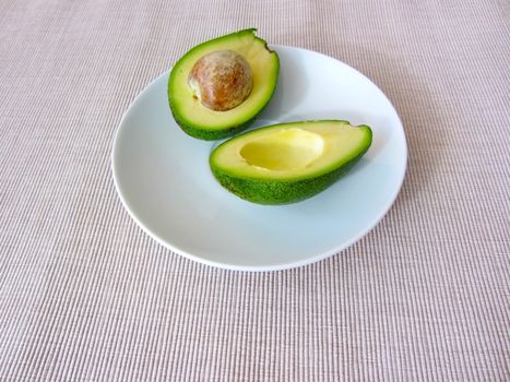 Fresh organic avocado halves, one with a bone, lie on a white plate on a grey cotton striped textured canvas. Healthy food concept.