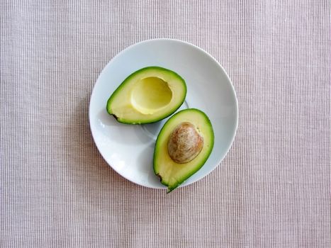 Fresh organic avocado halves, one with a bone, lie on a white plate on a grey cotton striped textured canvas, flat lay. Healthy food concept..