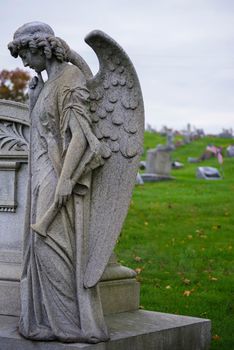 Sad angel with big wings and a trumpet stands off center with head bowed by a grave. Natural light with copy space, defocused background
