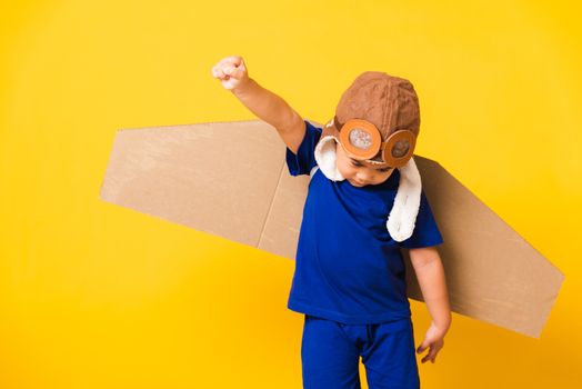 Happy Asian handsome funny child or kid little boy smile wear pilot hat play and goggles raise hand up with toy cardboard airplane wings flying, studio shot isolated yellow background, Startup freedom