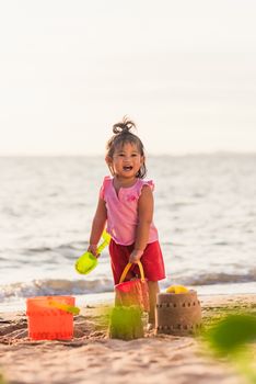 Happy fun Asian child cute little girl playing sand with toy sand tools at a tropical sea beach in holiday summer on sunset time, tourist trip concept